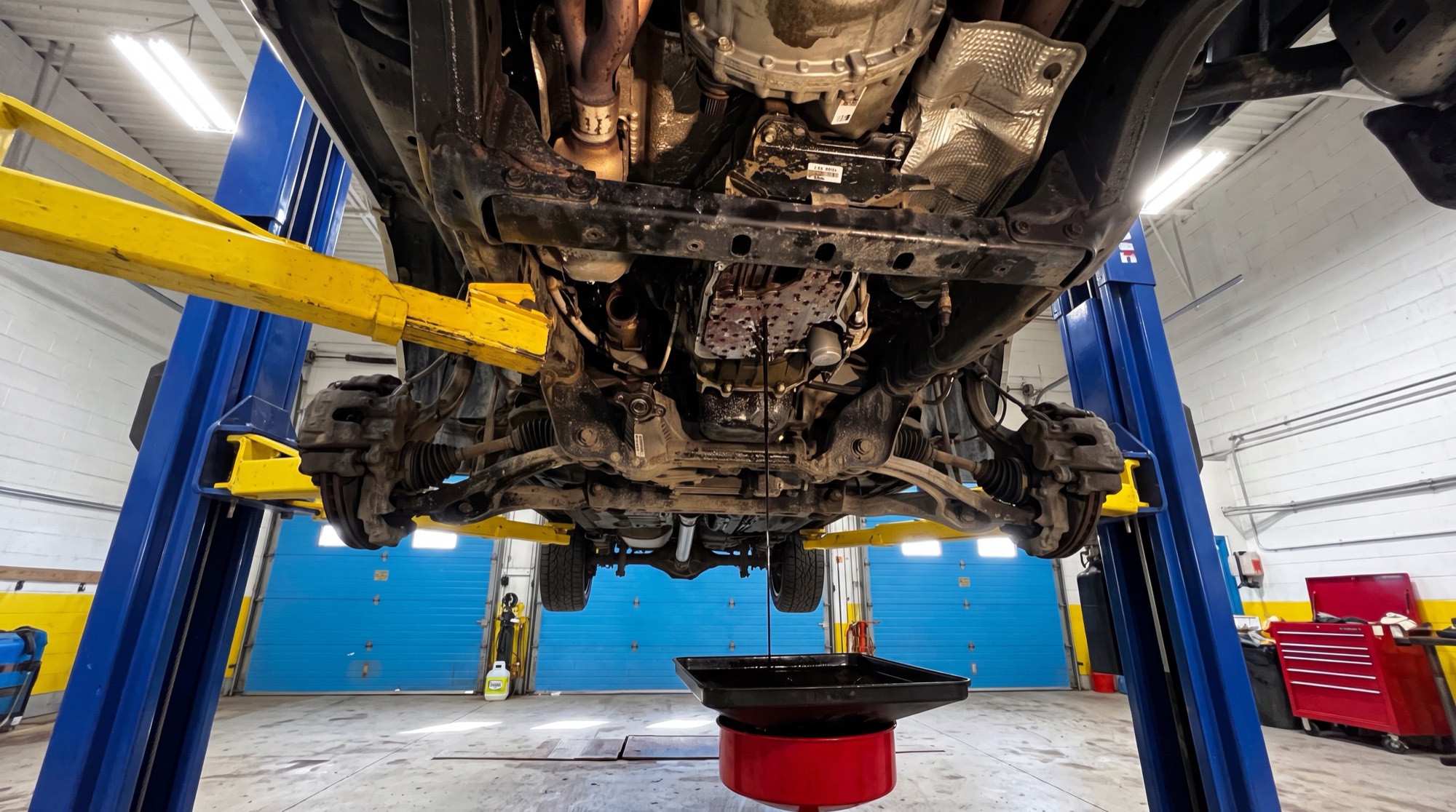 The underside of a vehicle on the lift, oil draining into a pan, exhaust and heat shield visible.
